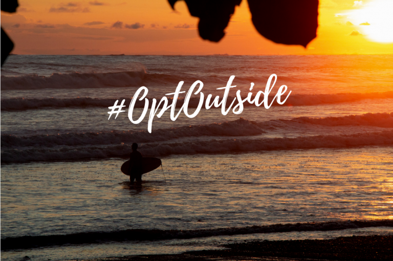 Surfer holding a surfboard in the ocean at sunset with text #OptOutside