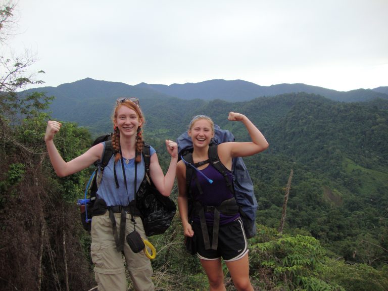 Two girls in the mountains wearing backpacks flexing their biceps