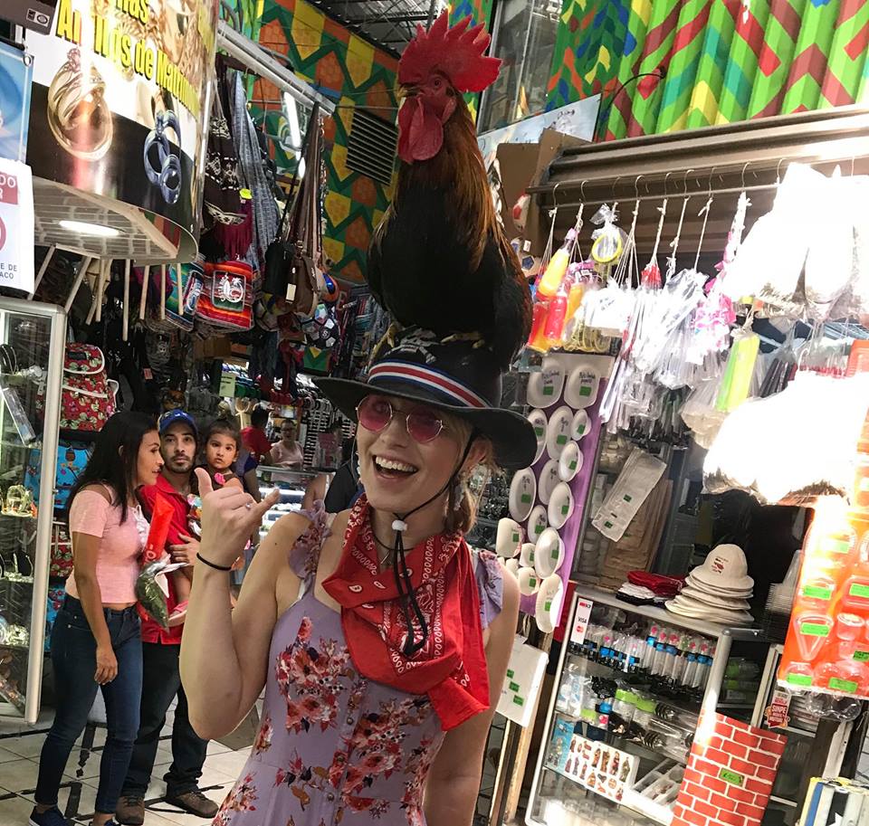 A girl wearing sunglasses with a rooster on her head in a market