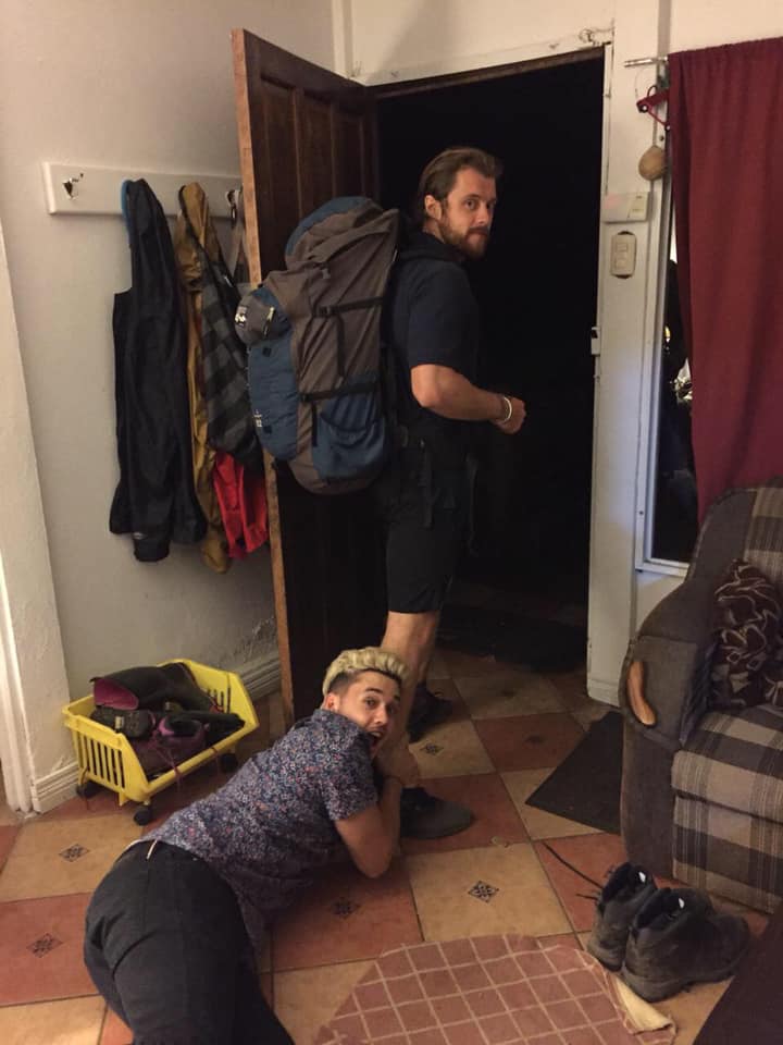 One man holding another man's leg as he is walking out a door with a backpack